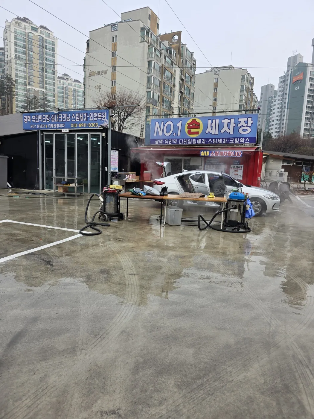 넘버원 손세차장 간판 아래에서 흰 승용차 문을 열어 두고 실내 스팀세차를 하는 모습